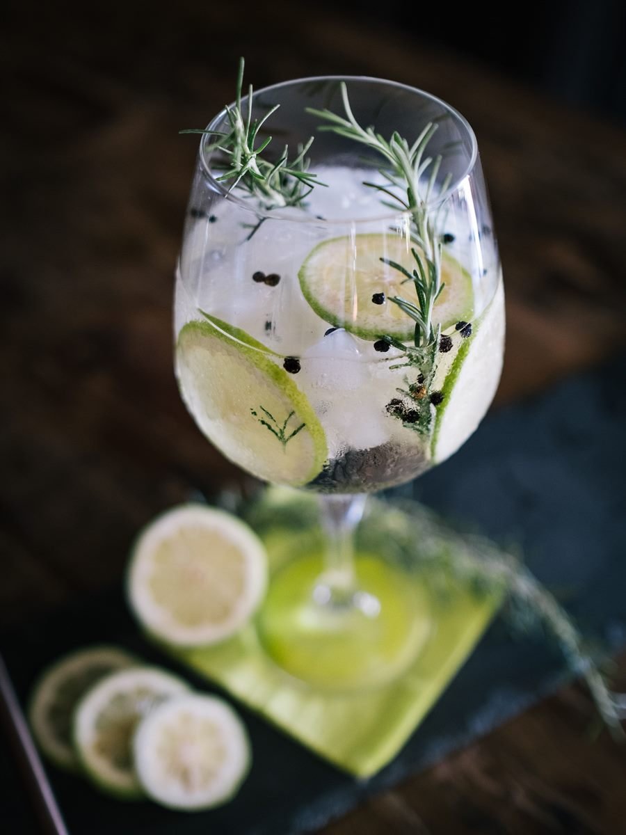 Gin tonic con romero, rodajas de limón y pimienta sobre madera oscura